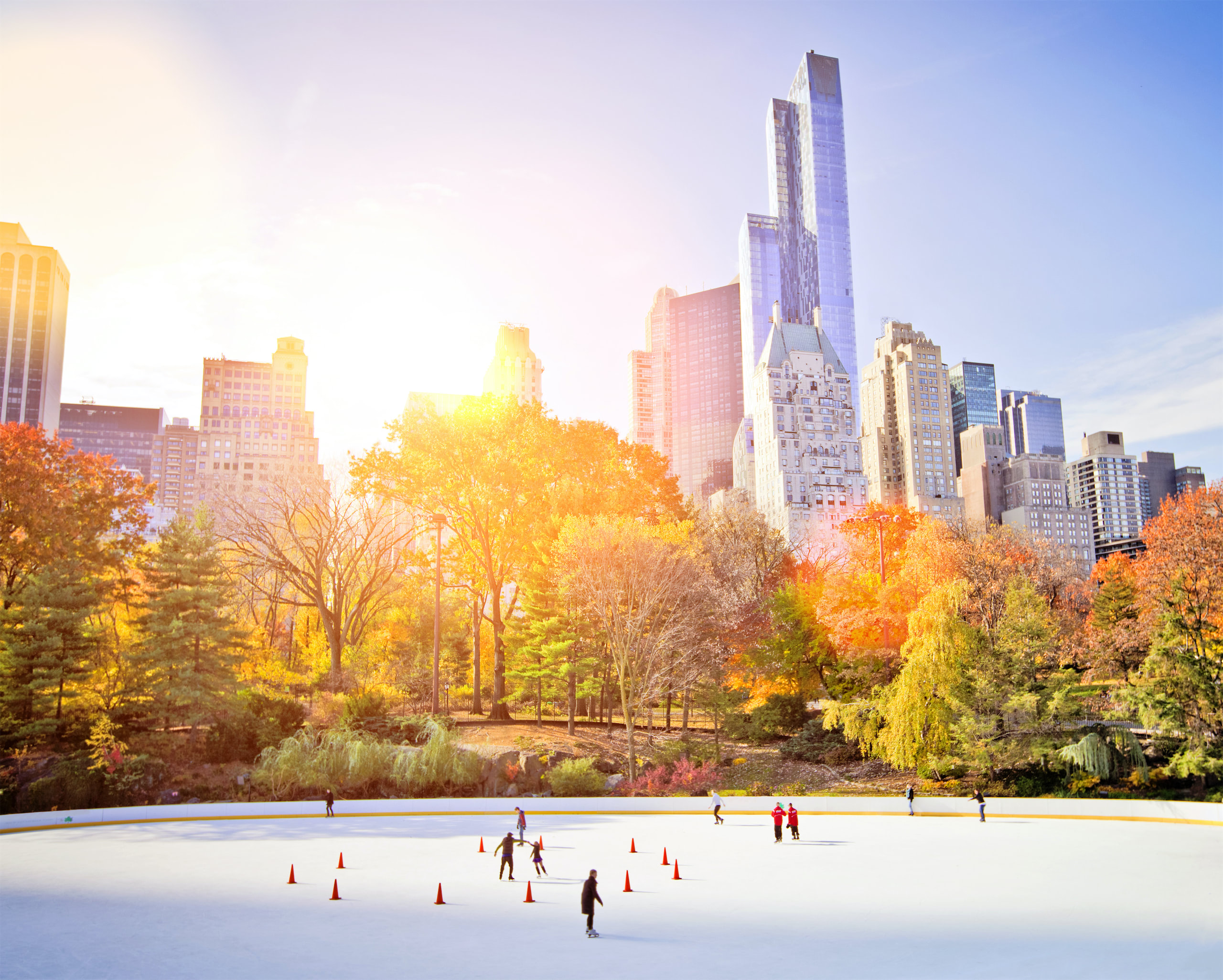 Skaters on Wollman Rink in autumn sunlight, with fall foliage and the Central Park South skyline behind