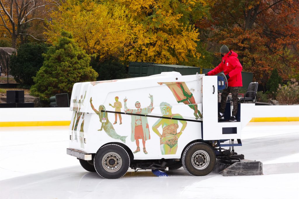 A Zamboni painted with an artist mural resurfacing the ice at Wollman Rink, autumn trees behind