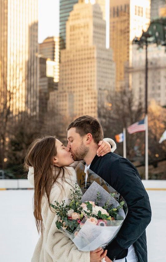 A newly engaged couple kissing on the ice at Wollman Rink, holding a bouquet, with Midtown towers behind them