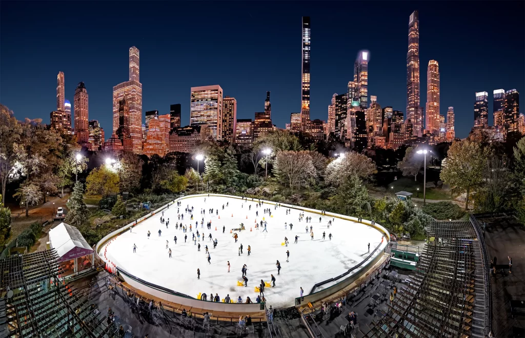 Aerial view of Wollman Rink at night, packed with skaters below the lit Central Park South skyline