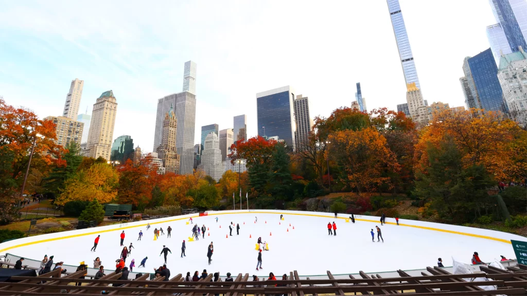 Skaters across the full Wollman Rink on an autumn day, with fall foliage and the Midtown skyline beyond