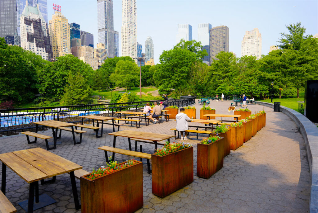 The Overlook terrace above Wollman Rink, set with wooden picnic tables and planters, with Central Park trees and the skyline beyond